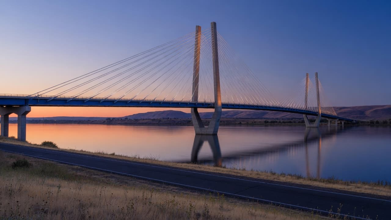 The Cable Bridge over the Columbia between Kennewick and Pasco at dusk, its white cables lit by the last light, dry grass along the riverside path.