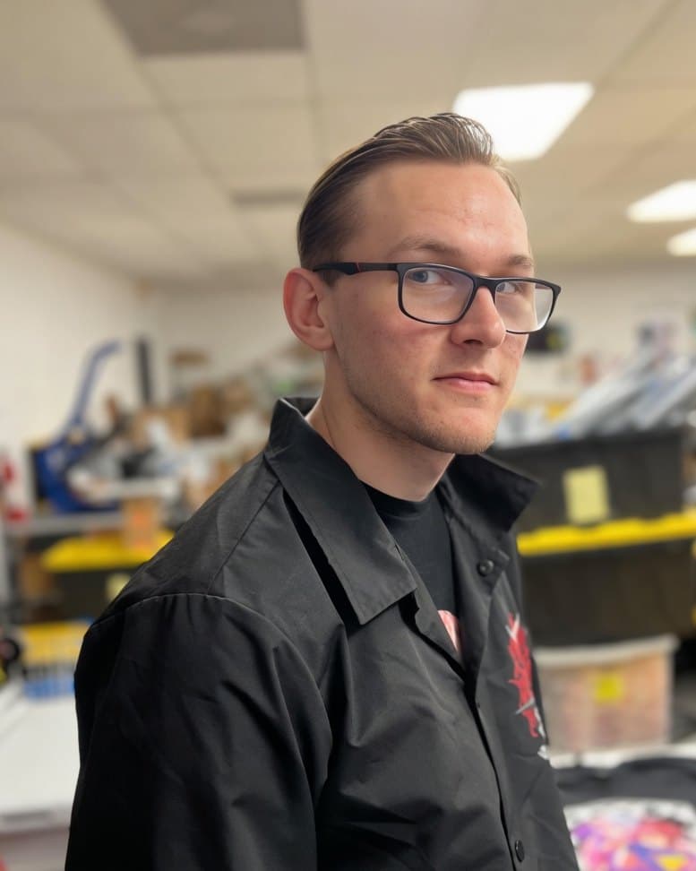Zack Grogan in a black work shirt and glasses, standing in a print shop.